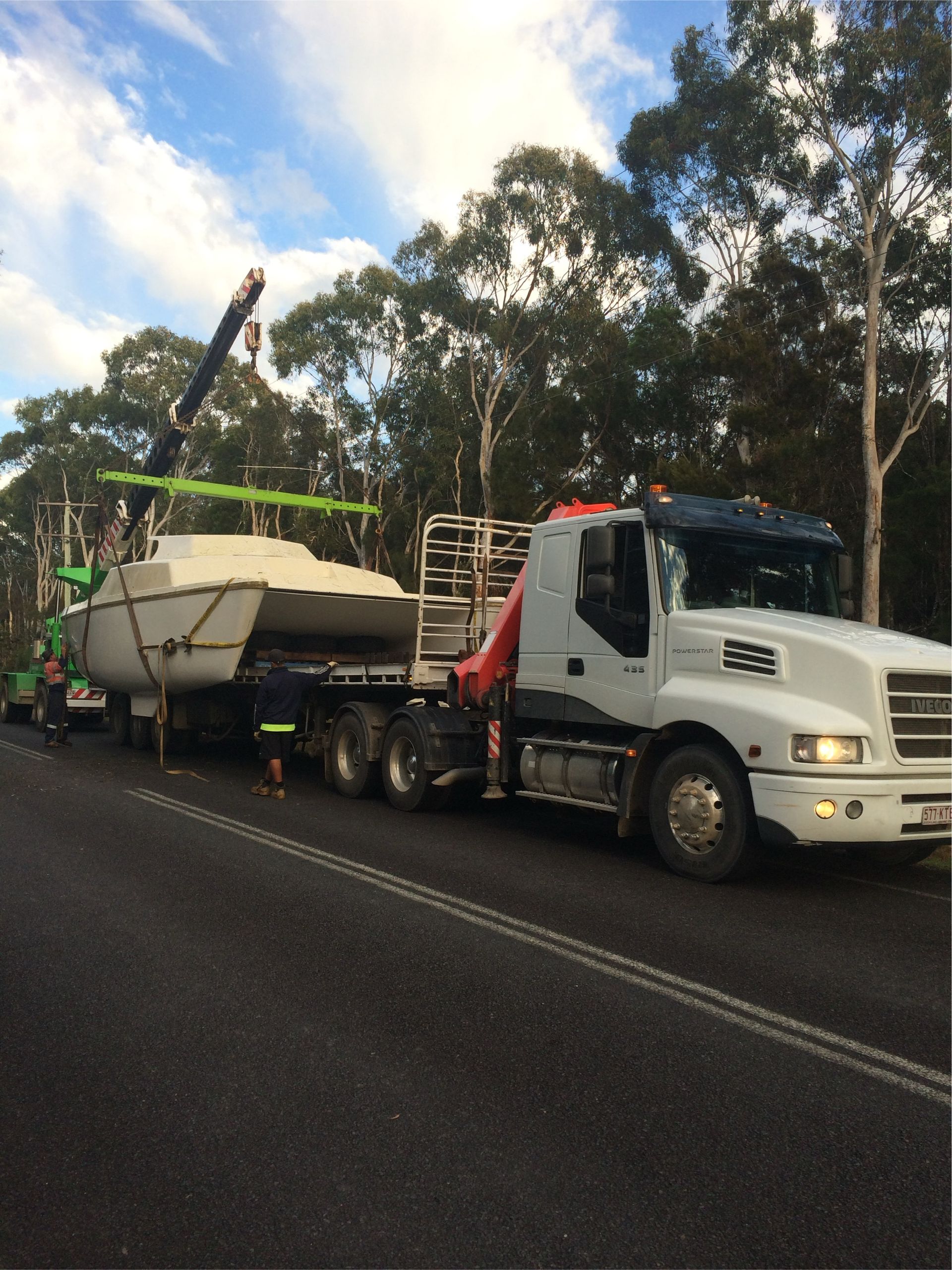Tow truck hauling a white boat on a highway beside trees under a cloudy sky — Trucking Crane Trucks in Warana, QLD
