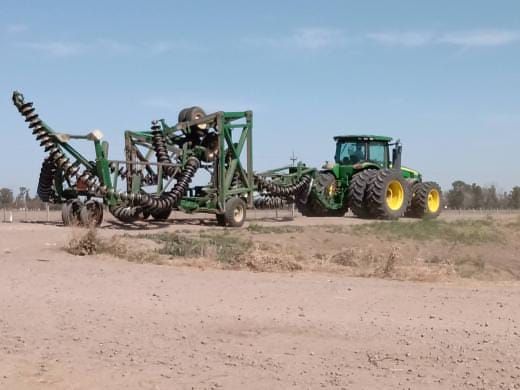 Un tractor verde tirando de una grada de discos agrícola, con un lado del equipo plegado, en un campo de tierra.