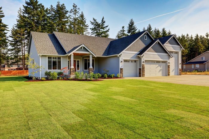 Single-story suburban house with a large green lawn, gray roof, and three-car garage under a clear sky