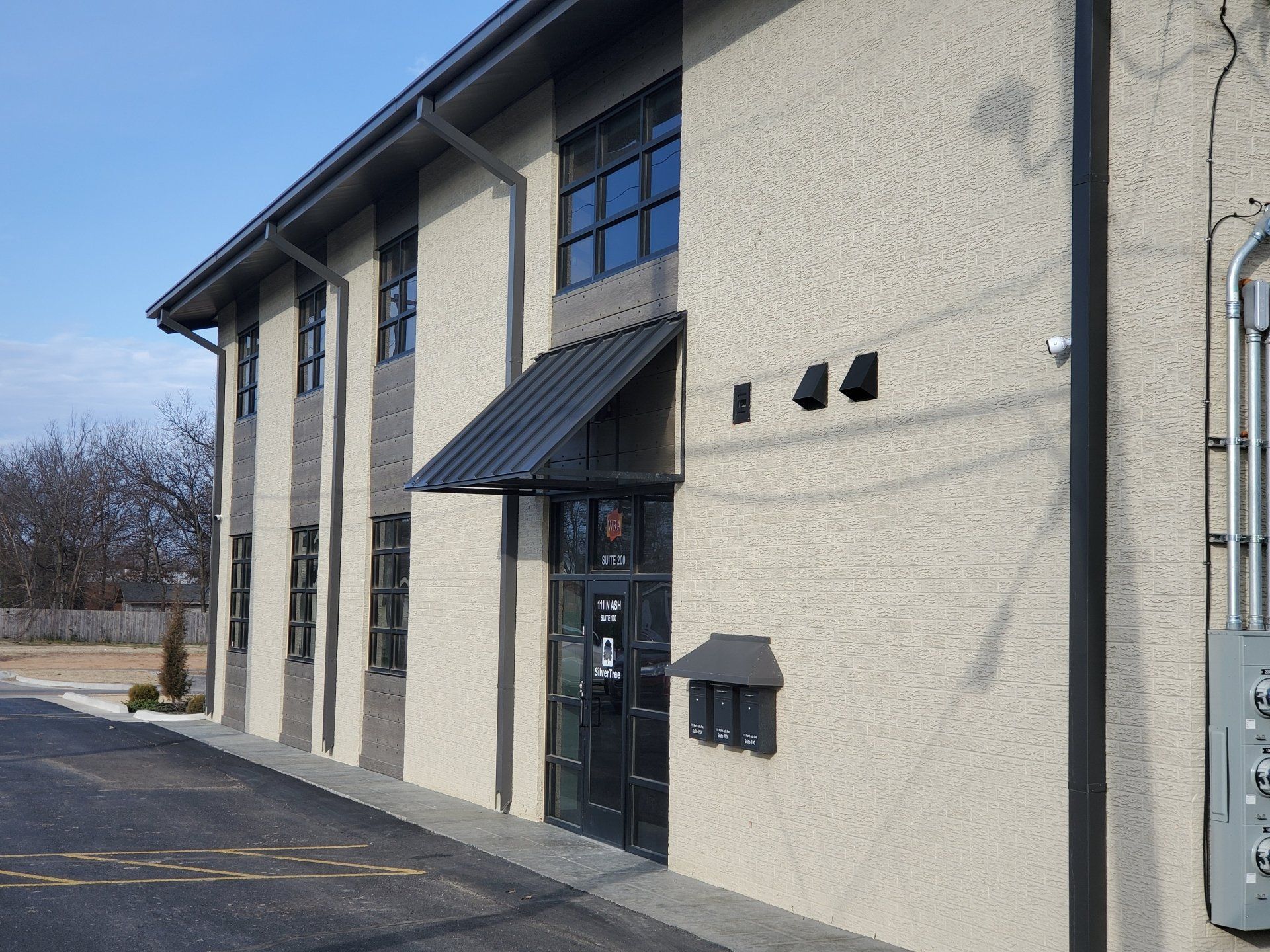 Exterior of a beige two-story building with a black awning and entrance door, beside a parking lot.