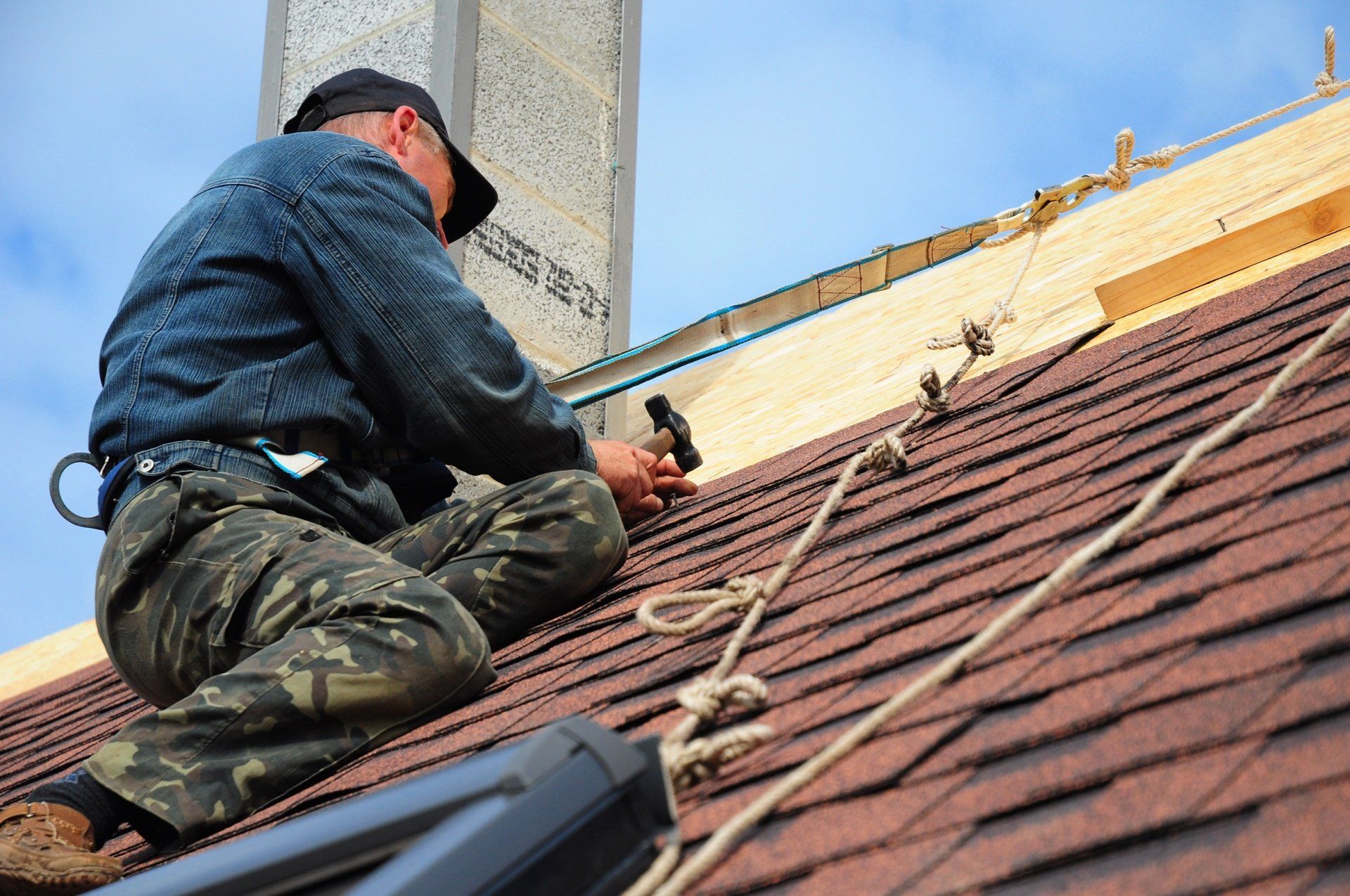 Roofer kneeling on a shingled roof, using a hammer near the roof ridge.