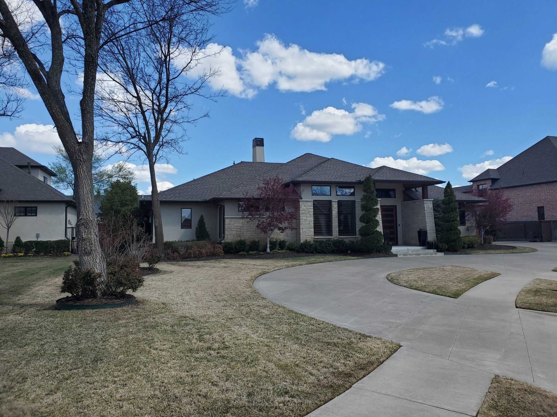 Suburban house with curved driveway, leafless tree, and partly cloudy blue sky.