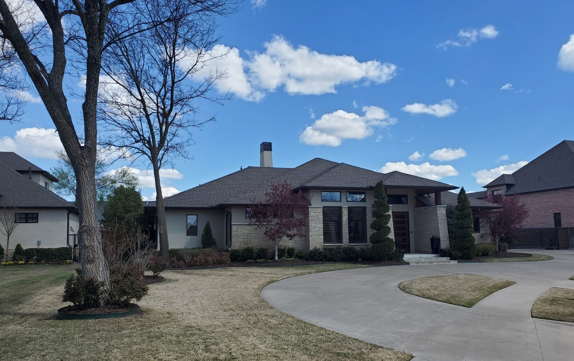 Suburban house with a curved driveway, leafless trees, and a blue sky with scattered clouds.