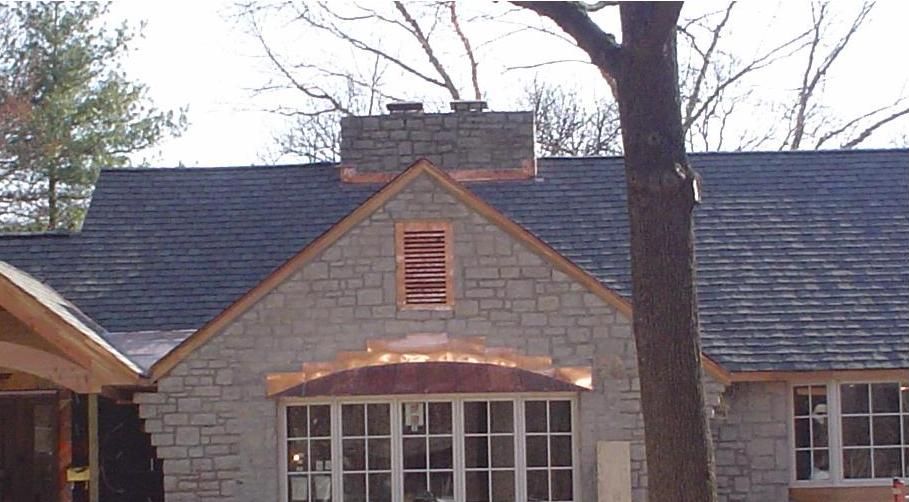 Stone house with dark roof, central gable, and large front window partially obscured by a tree trunk.