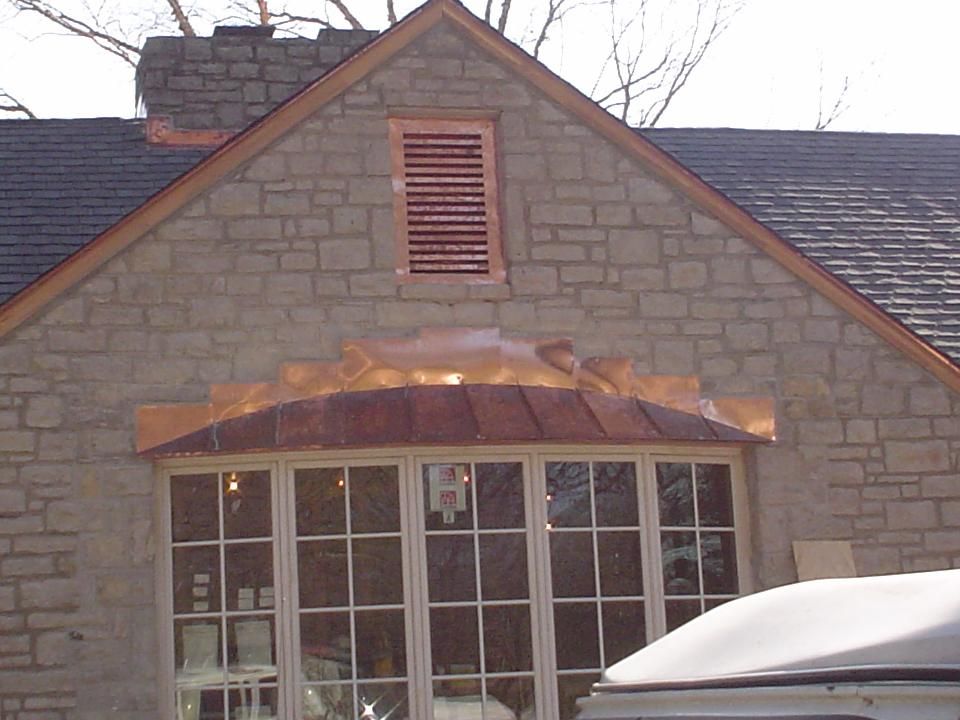 Stone house gable with a small louvered vent and copper awning over a window, roof and car visible