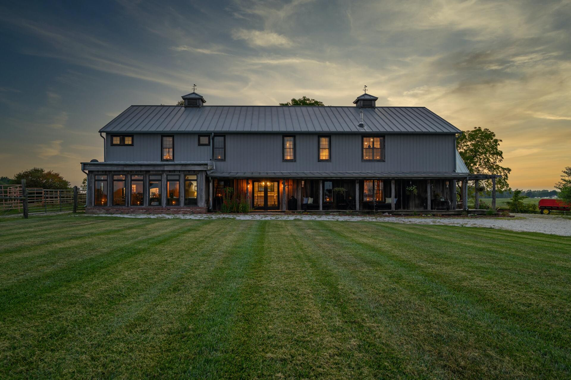 Large gray barn-style house at dusk with warm lights and a wide green lawn