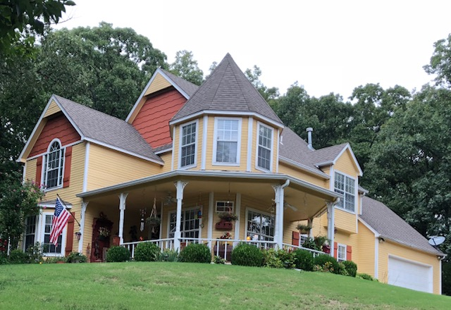 Yellow Victorian-style house with red trim, wraparound porch, and two-story turret on a grassy hill