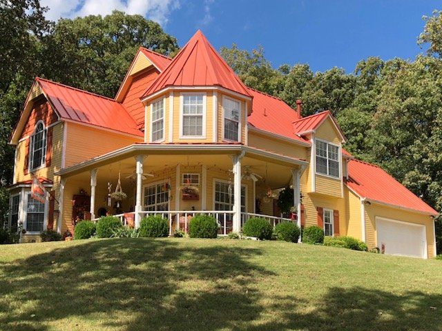 Large yellow house with red roof and wraparound porch on a grassy hillside under a blue sky