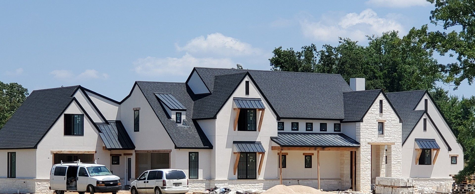 Modern stone-and-gray suburban house with multiple gables and parked vehicles under a blue sky