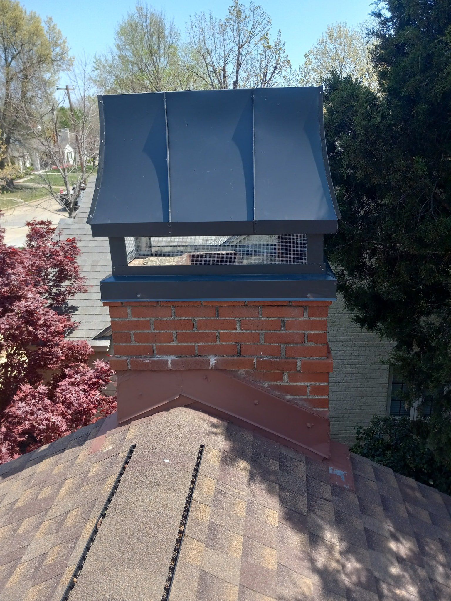 Brick chimney with a black metal cap above a shingled roof, surrounded by trees and shrubs.