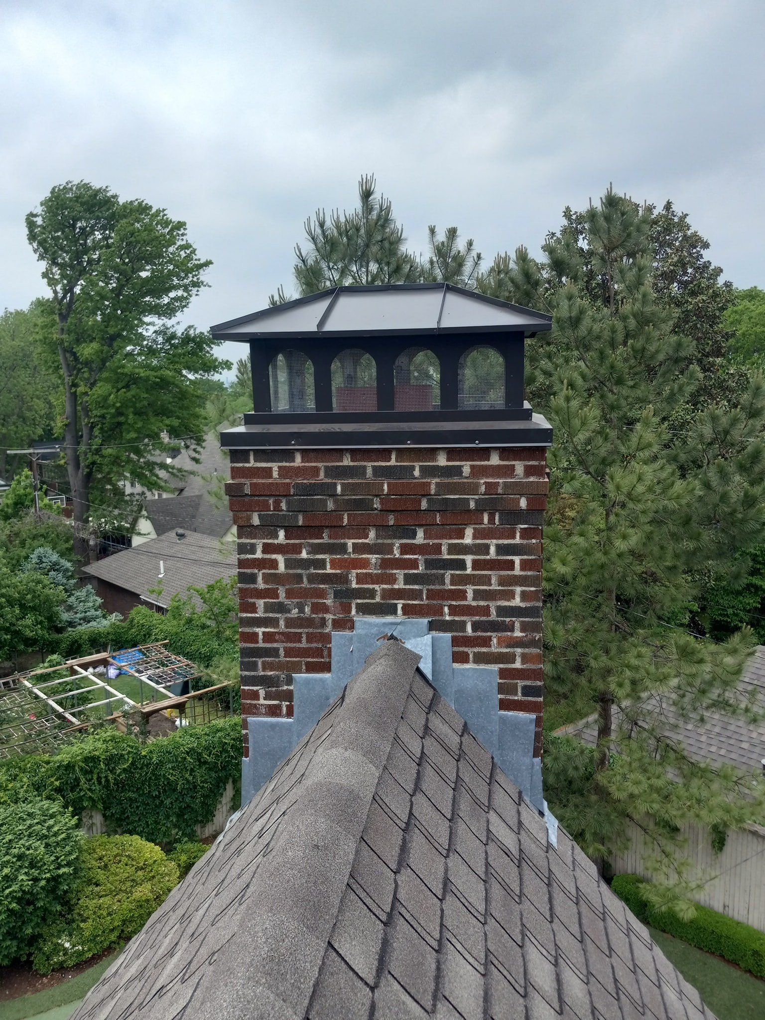 Brick chimney with black cap rising above a gray shingled roof, surrounded by trees and gardens