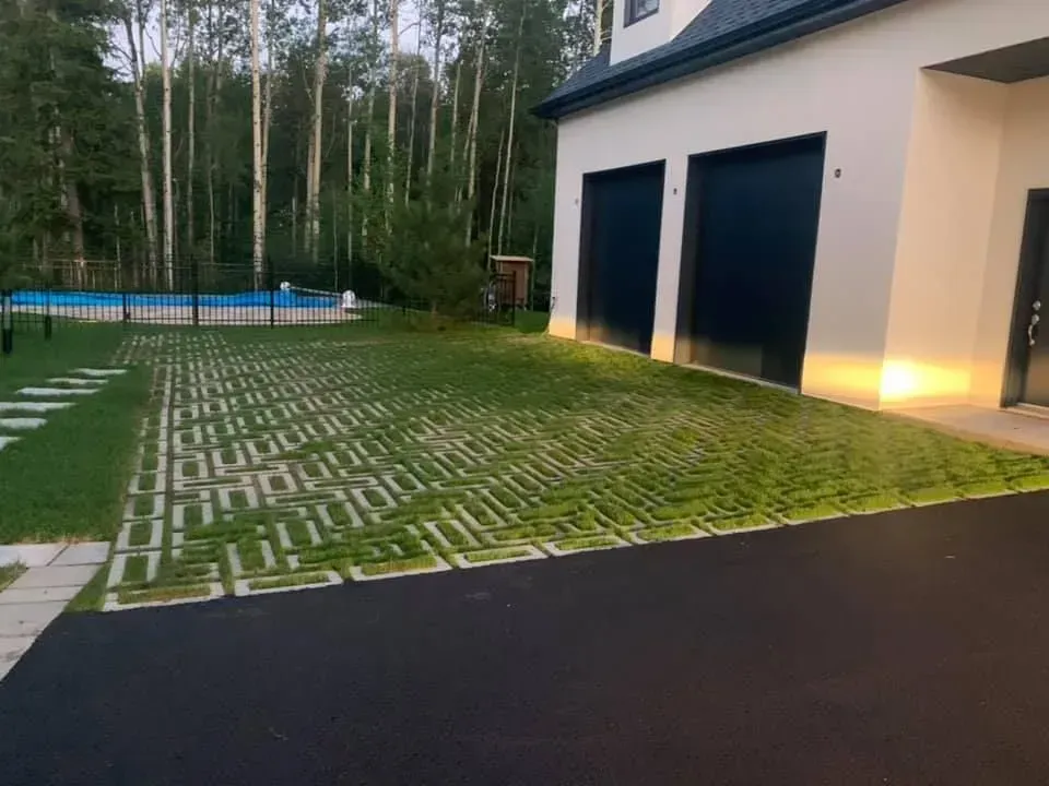 Maison moderne avec deux portes de garage sombres, une entrée latérale éclairée et une pelouse pavée bordée d'une piscine et d'arbres.