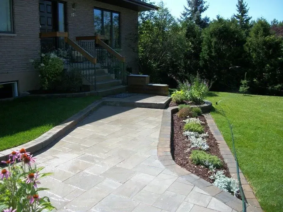 Allée en pierre incurvée longeant une maison, bordée de parterres de fleurs et d'une pelouse verdoyante baignée de soleil.