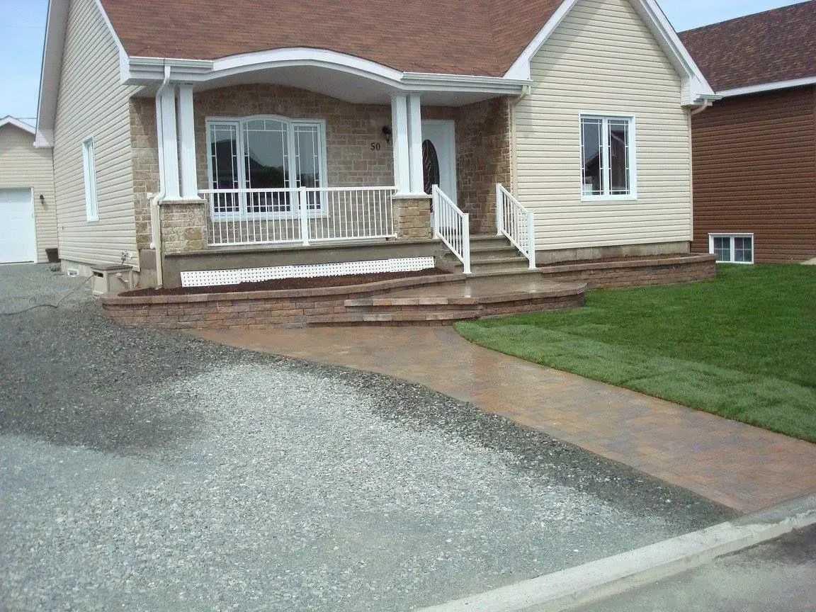 Vue de face d'une maison beige avec un porche blanc, une allée en gravier et un trottoir incurvé.