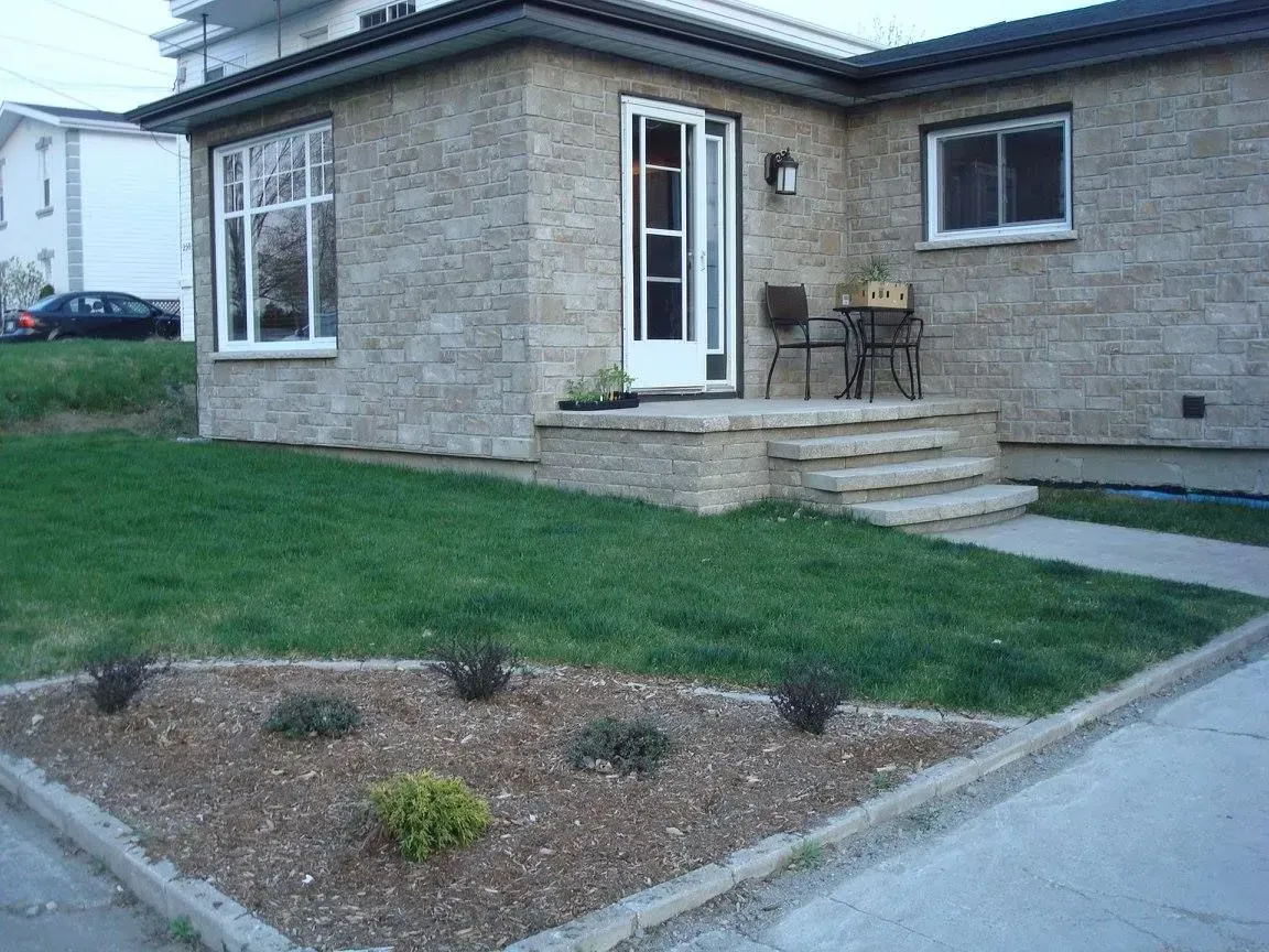 Maison en briques avec marches d'entrée, chaises de patio et un petit parterre de gravier aménagé à côté d'une pelouse.