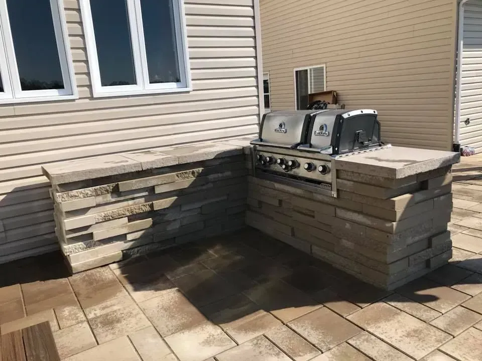Terrasse extérieure avec bancs en pierre intégrés et barbecue en acier inoxydable adossé à un mur beige.