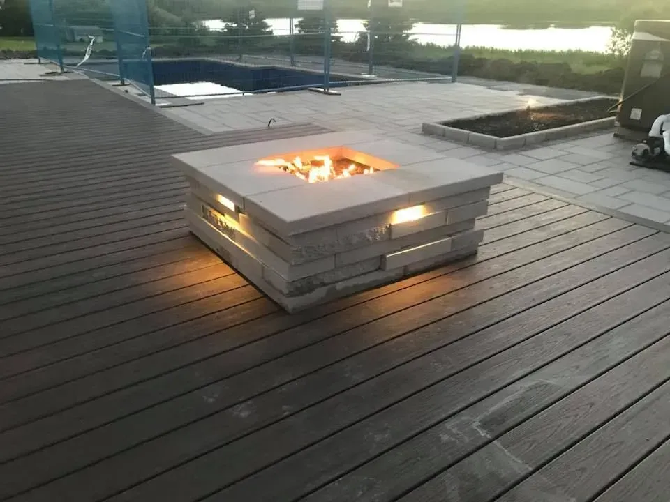 Foyer carré en métal illuminé sur une terrasse en bois au coucher du soleil, avec piscine et sièges en arrière-plan
