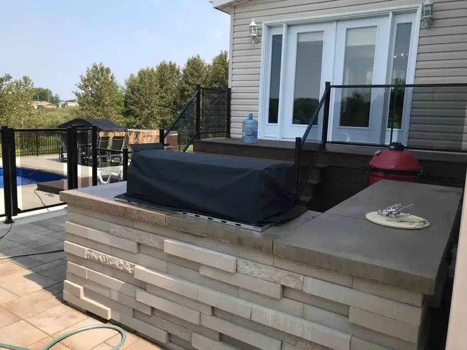 Terrasse avec barbecue couvert, comptoir en pierre et balustrade en verre, jouxtant une maison et une piscine.