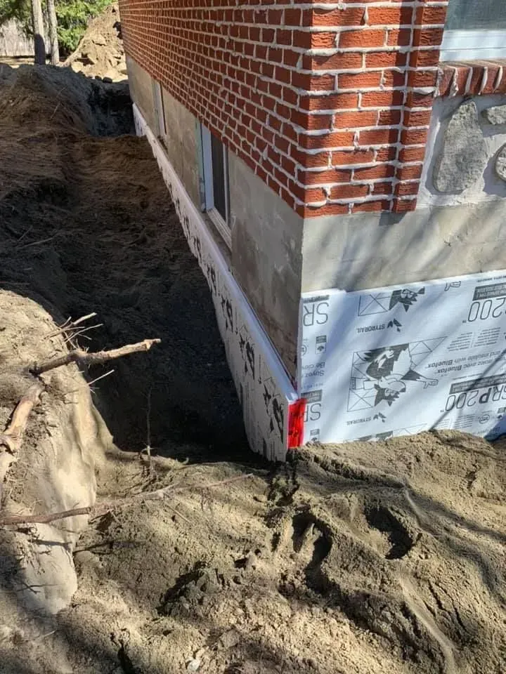 Fondations de maison en briques recouvertes d'une membrane d'étanchéité, avec une tranchée de terre apparente sur le côté.