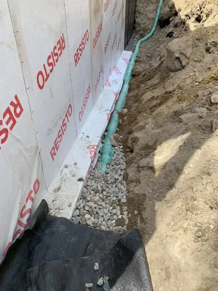 Mur de fondation recouvert d'isolant blanc à côté d'une tranchée remplie de gravier et d'un tuyau d'arrosage vert.