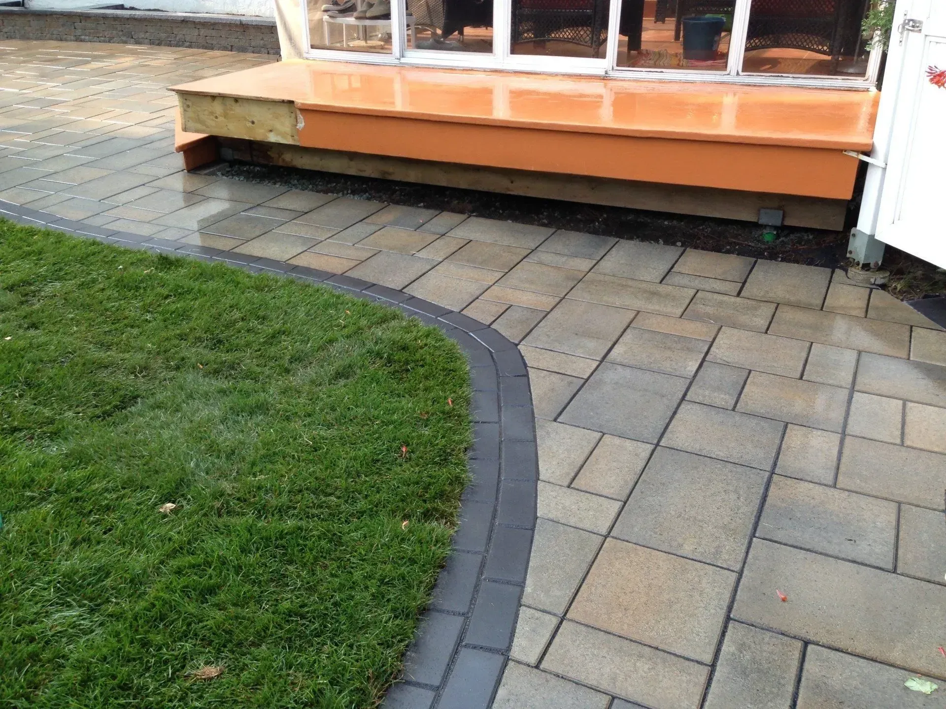 Allée pavée incurvée longeant une pelouse et une terrasse en bois orange avec fenêtres.