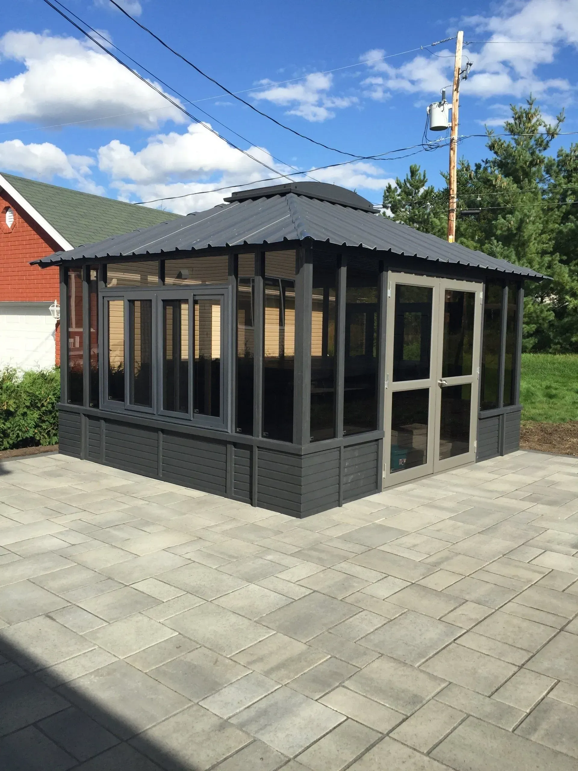 Petit pavillon aux parois de verre gris foncé sur une terrasse pavée sous un ciel partiellement nuageux