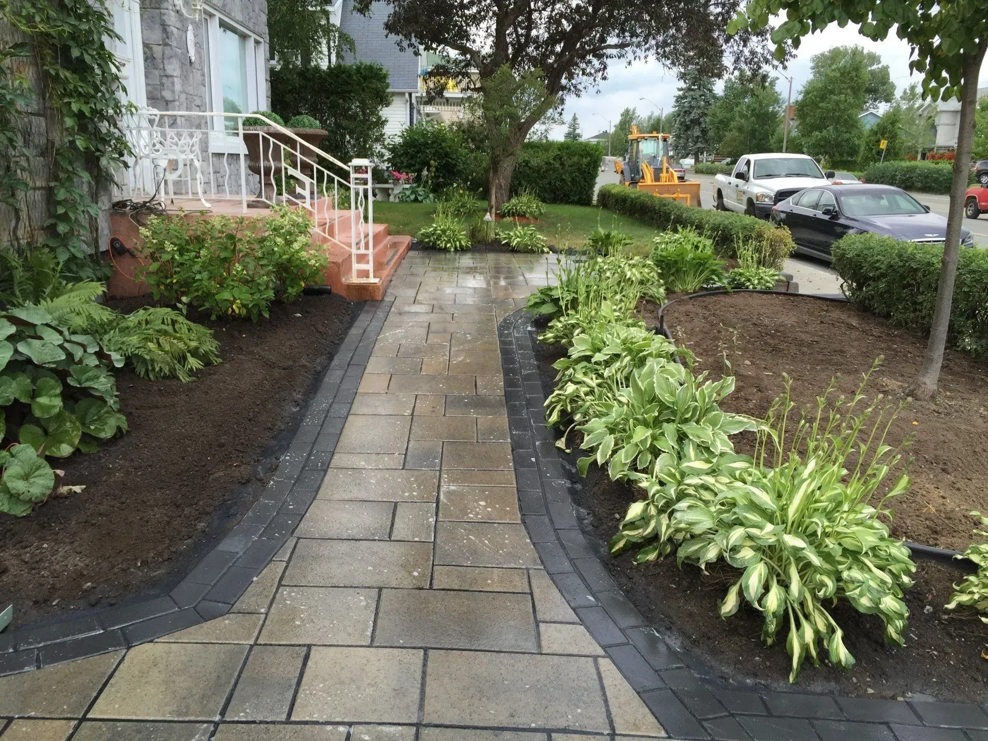 Allée de jardin détrempée par la pluie, pavée de pierres, bordée de plantes luxuriantes et de voitures garées le long d'une rue résidentielle