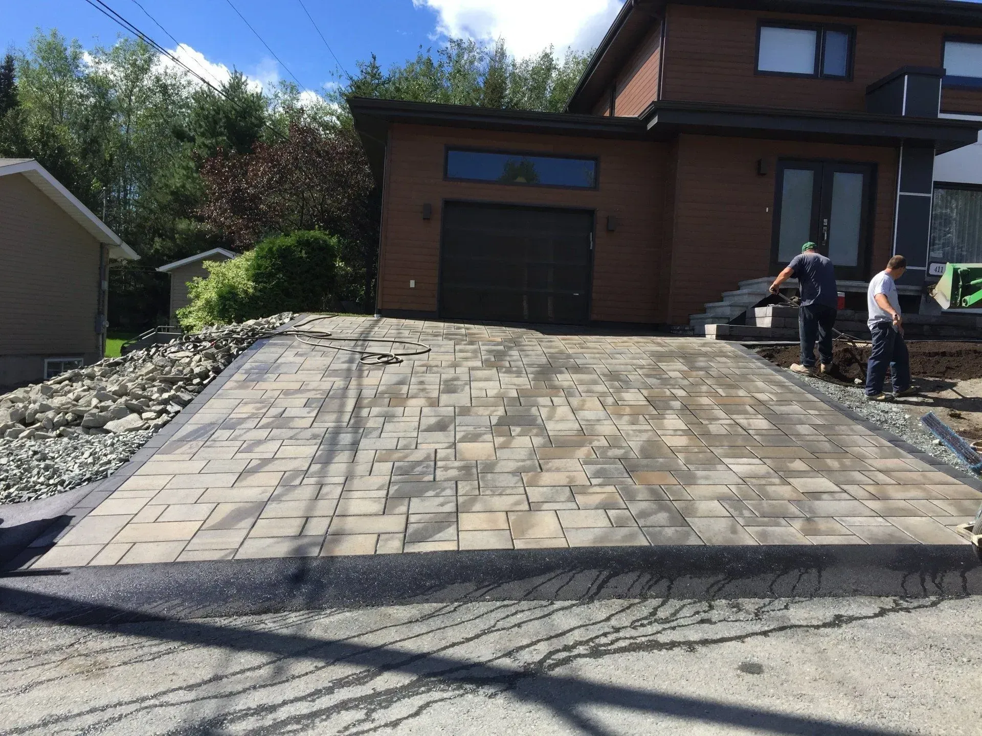 Deux ouvriers pavant une allée en pente devant une maison brune avec un garage