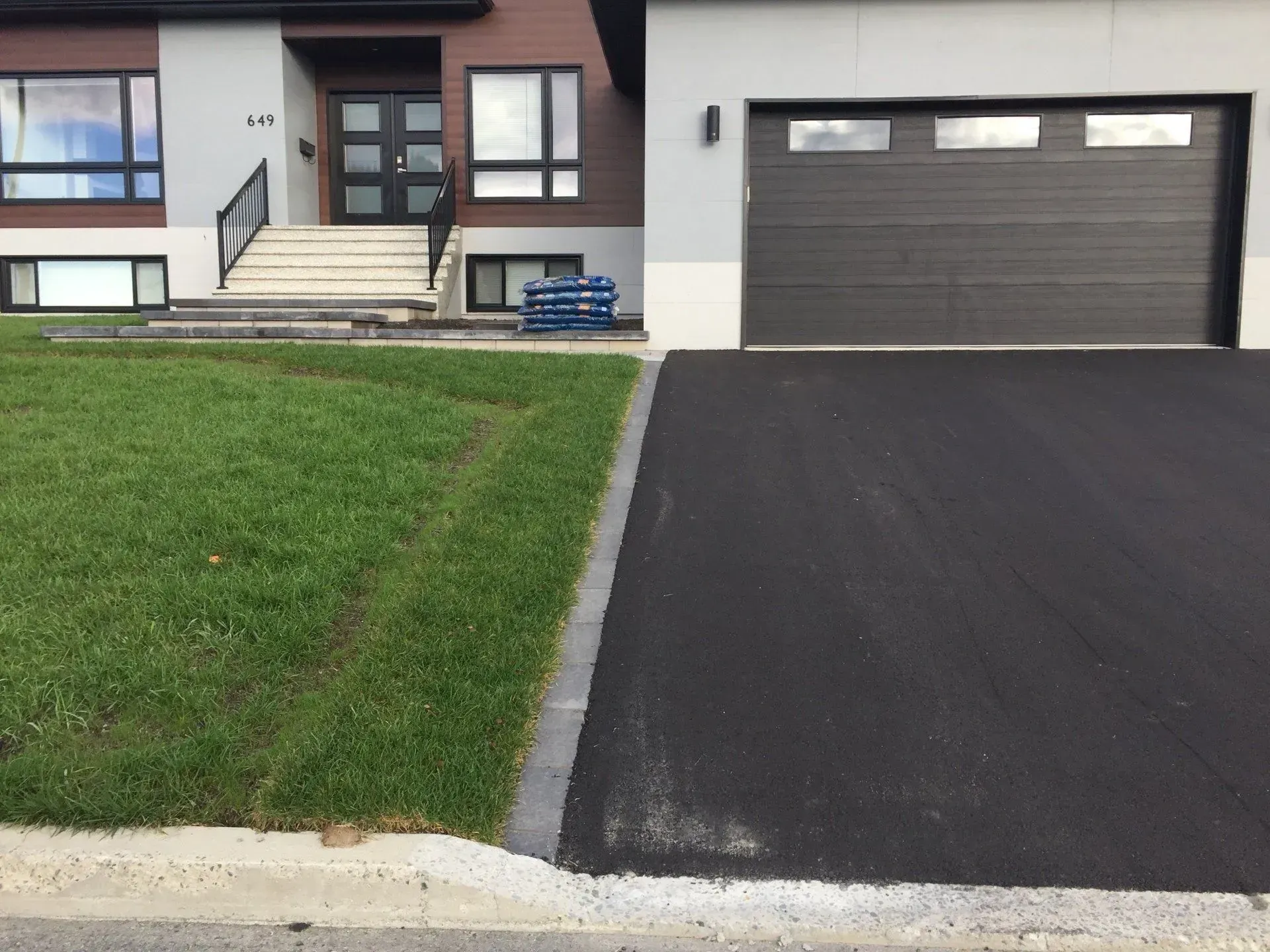 Maison moderne avec une porte de garage foncée, une allée noire et une pelouse verte devant.