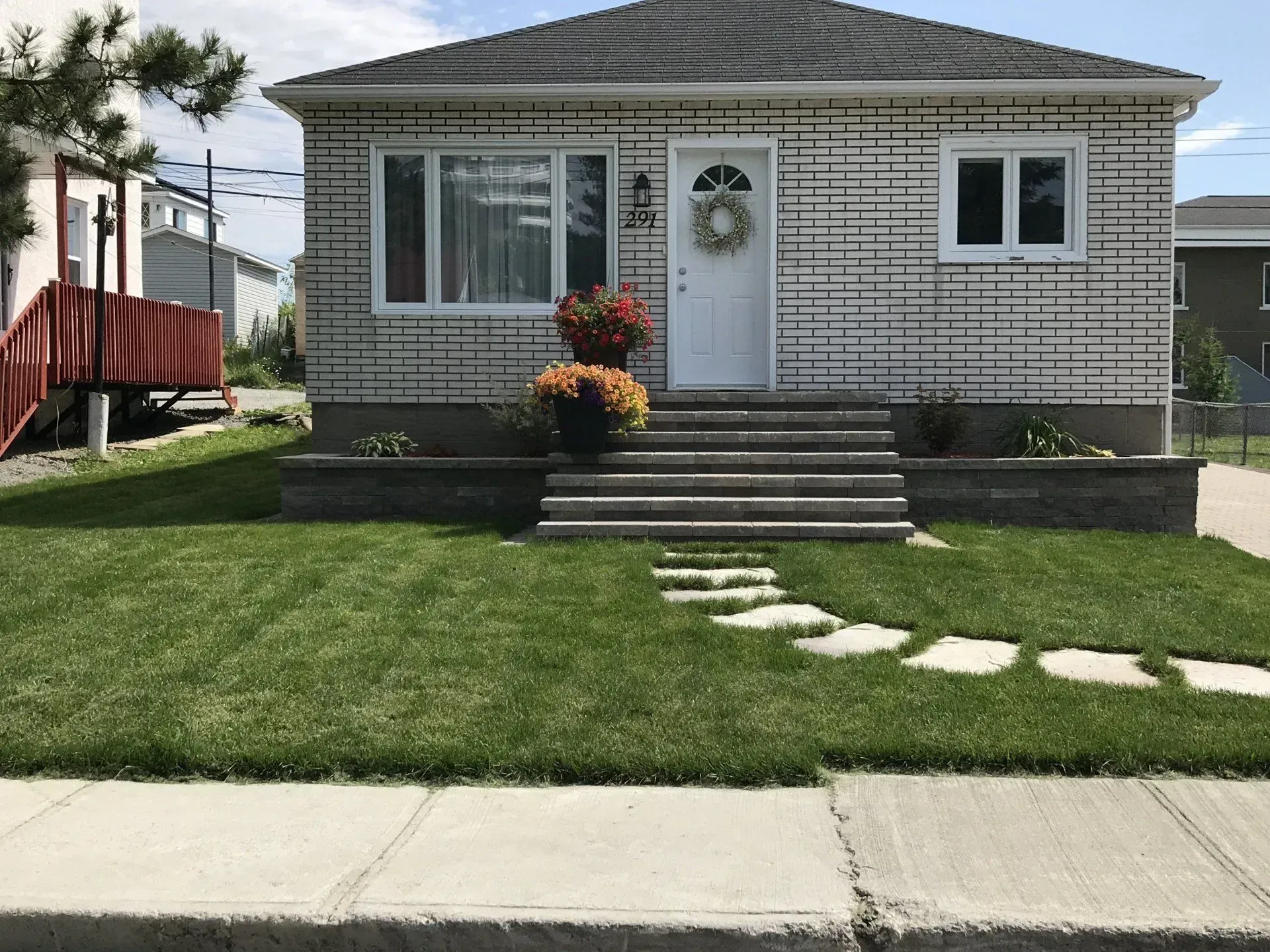 Maison beige de plain-pied avec un perron, une porte blanche et une pelouse verte bordant un trottoir.
