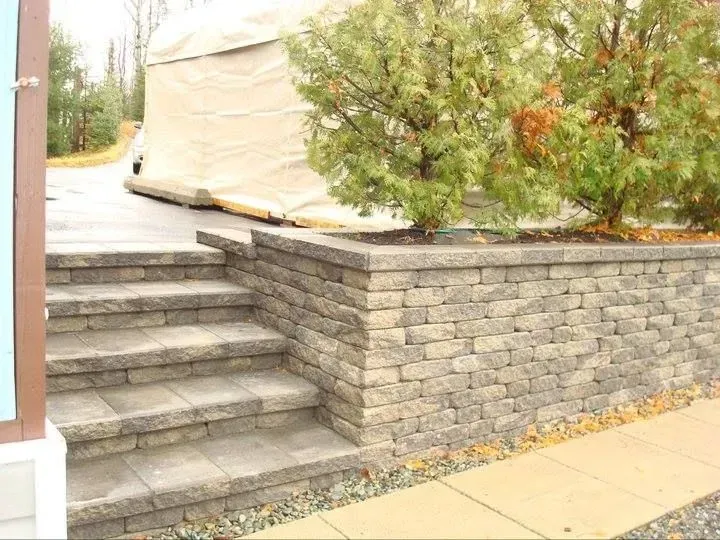 Des marches en pierre longent un mur de soutènement gris et un trottoir bordé d'arbustes, surmonté d'une structure couverte.