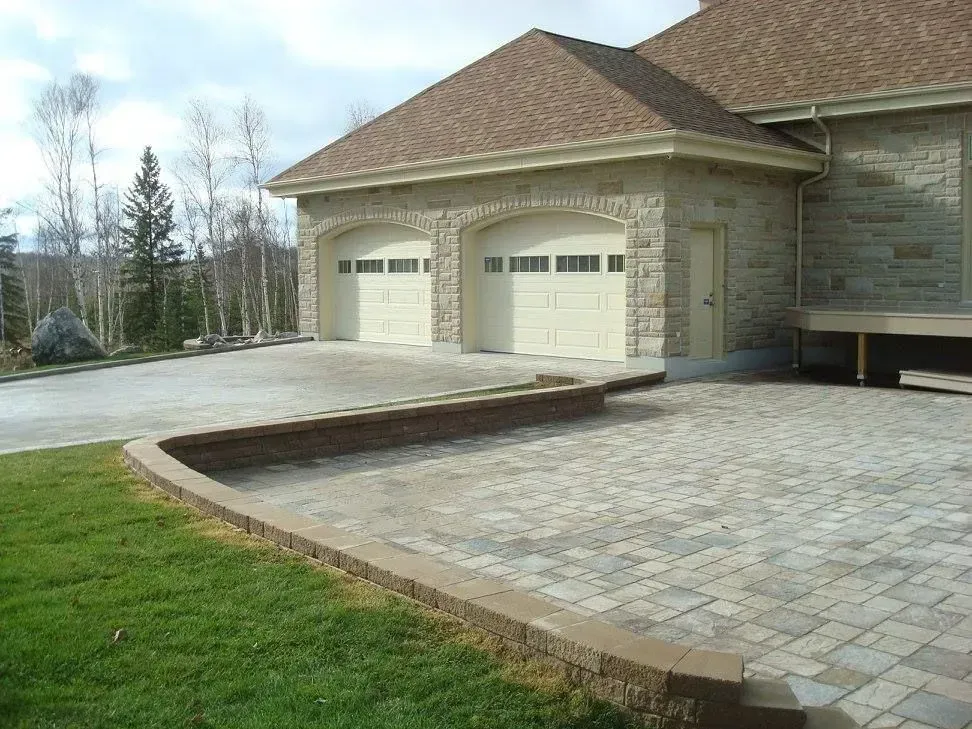 Maison en pierre avec deux portes de garage et une allée pavée donnant sur une pelouse verdoyante.