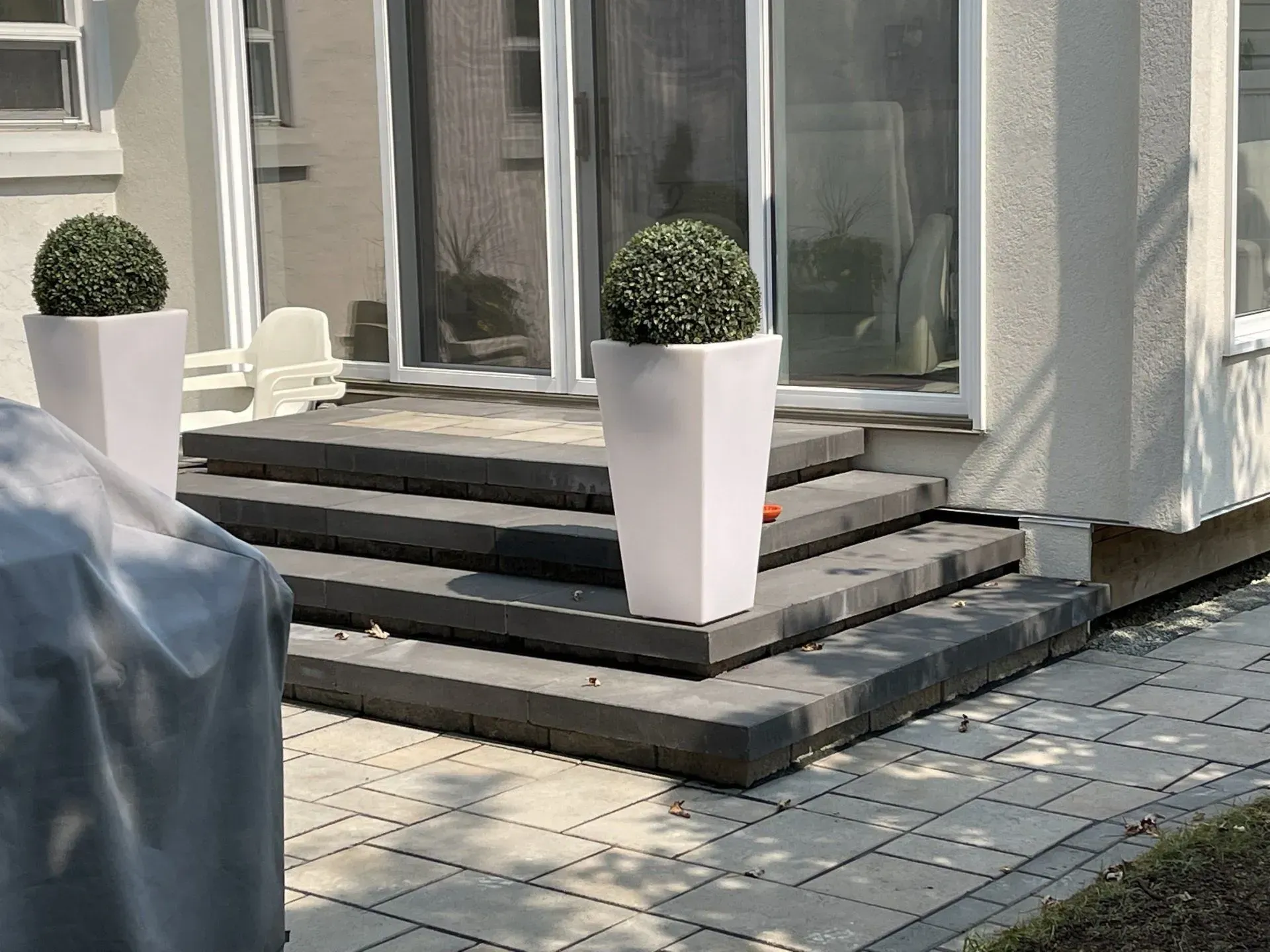 Entrée de maison moderne avec marches grises, deux jardinières blanches et un objet couvert sur la terrasse