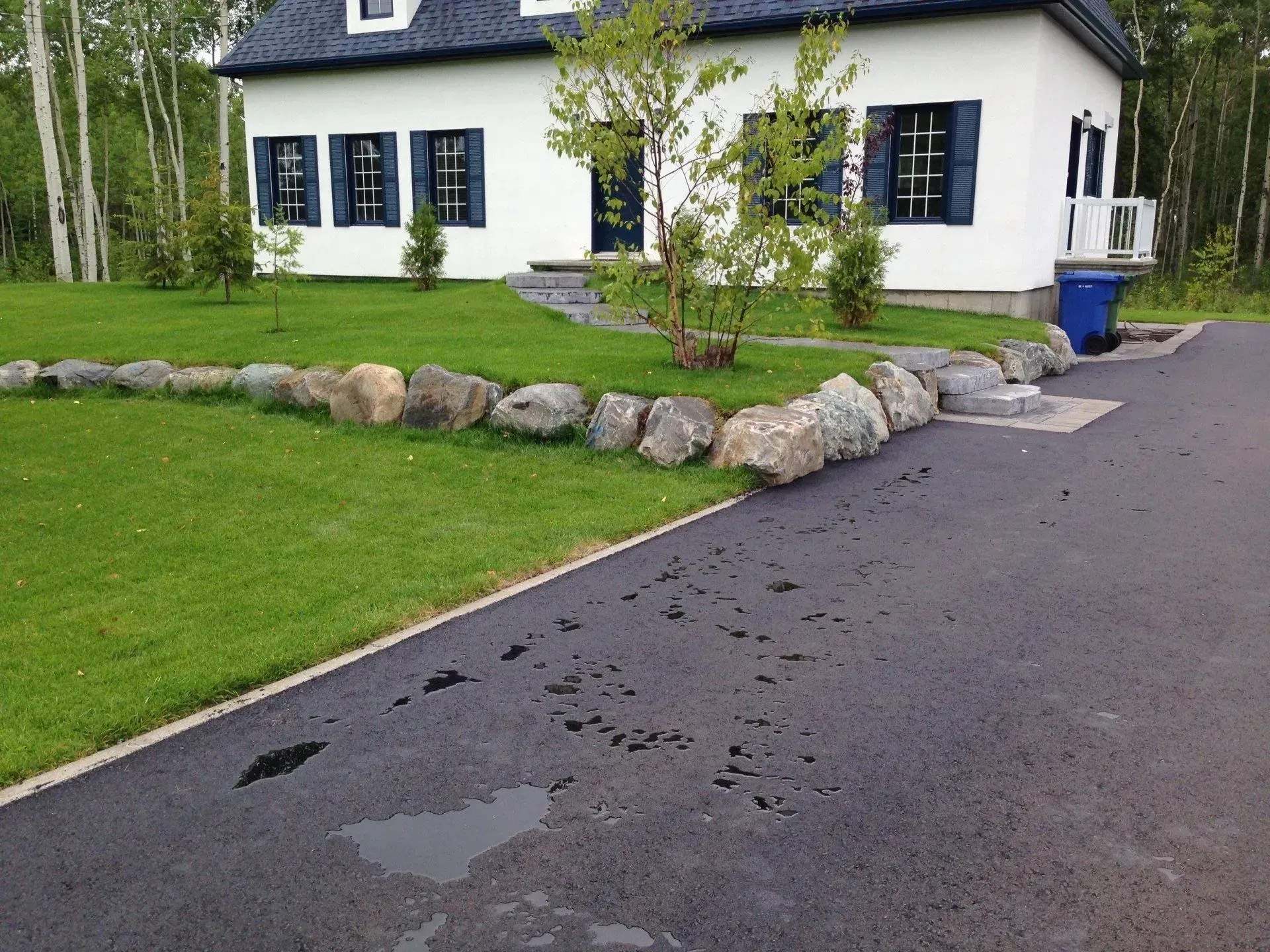 Maison blanche aux volets bleus, bordée d'une allée asphaltée mouillée et d'un jardin gazonné agrémenté de rochers et d'arbres.