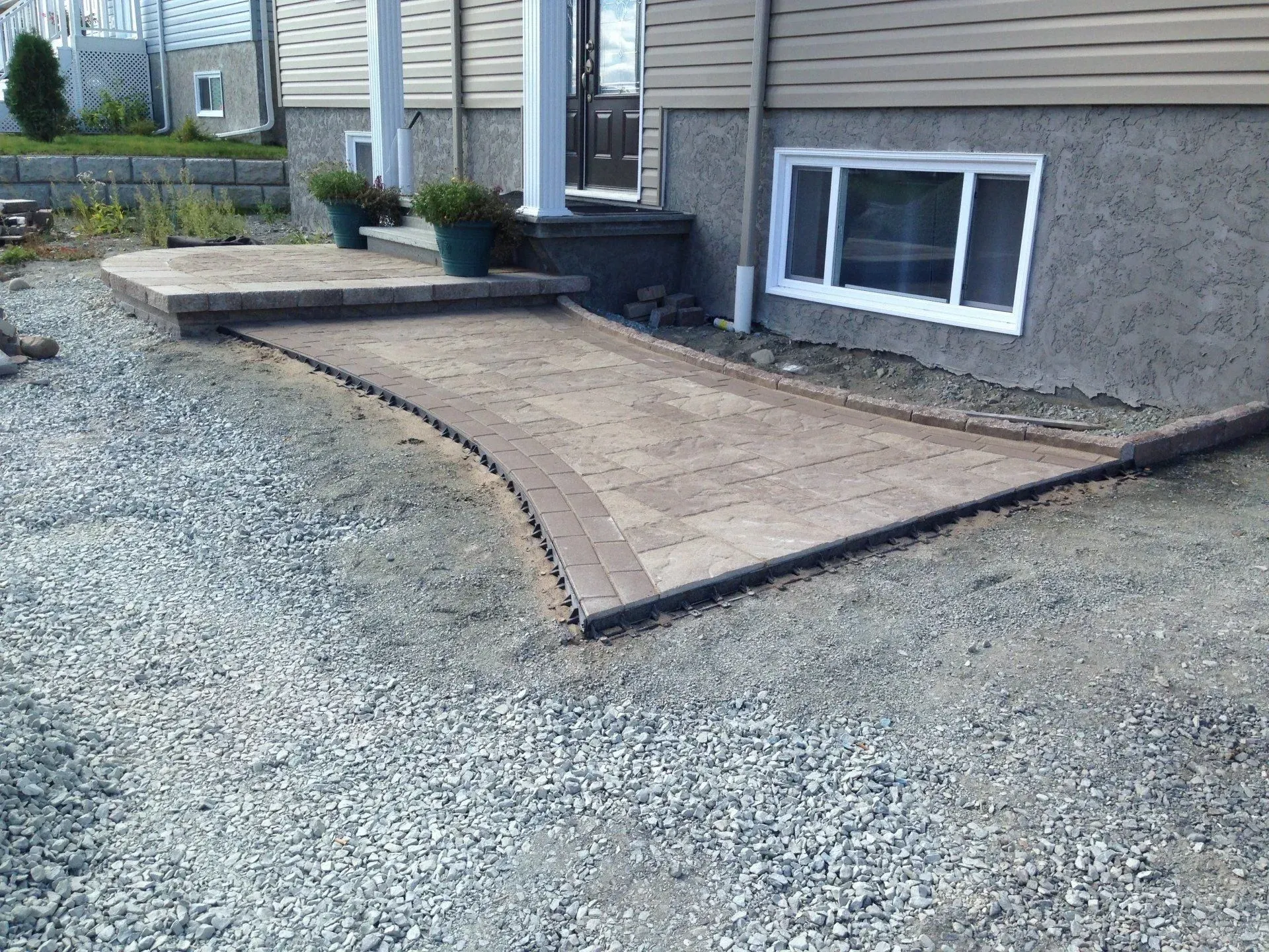 Allée et rampe en béton menant à l'entrée d'une maison, à côté d'une fenêtre de sous-sol, entourées de gravier.