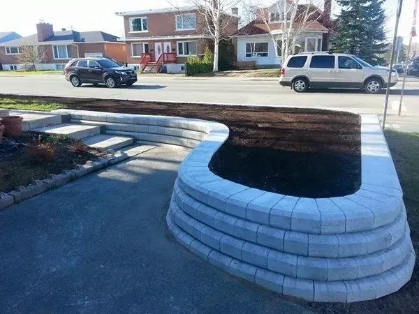 Jardinière incurvée en pierre blanche le long d'une allée pavée dans un quartier résidentiel, avec des maisons et des voitures garées à l'arrière.