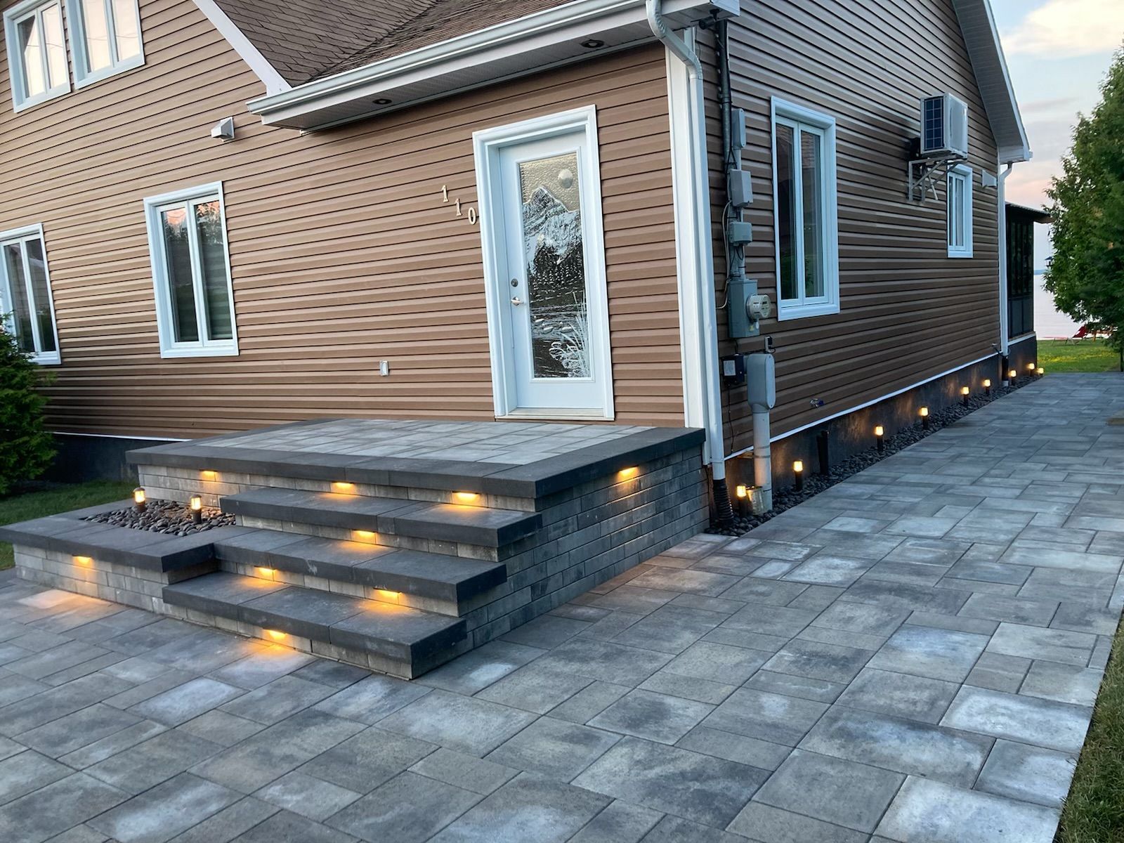 Maison moderne en briques avec un perron éclairé et une terrasse pavée au crépuscule