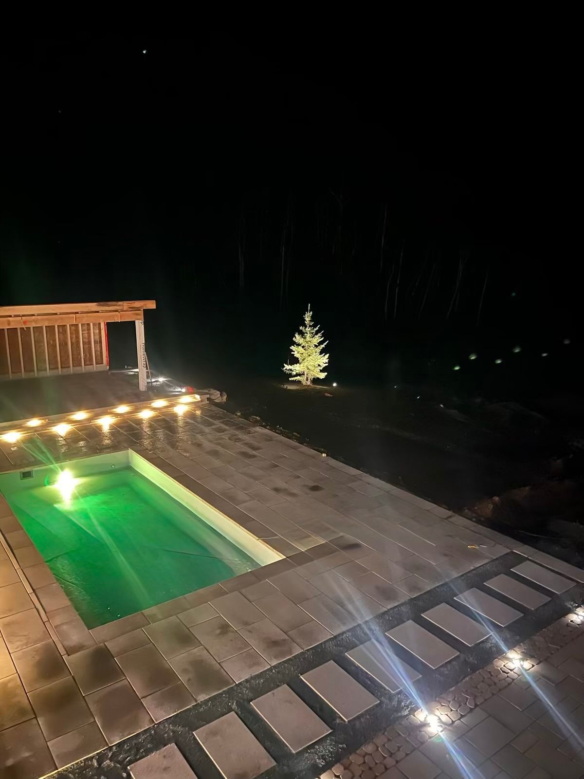 Terrasse nocturne avec piscine verte illuminée, guirlandes lumineuses et petit arbre illuminé au-delà du patio.