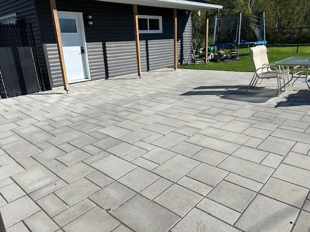Terrasse pavée jouxtant une maison grise, avec des chaises d'extérieur et une table en arrière-plan.