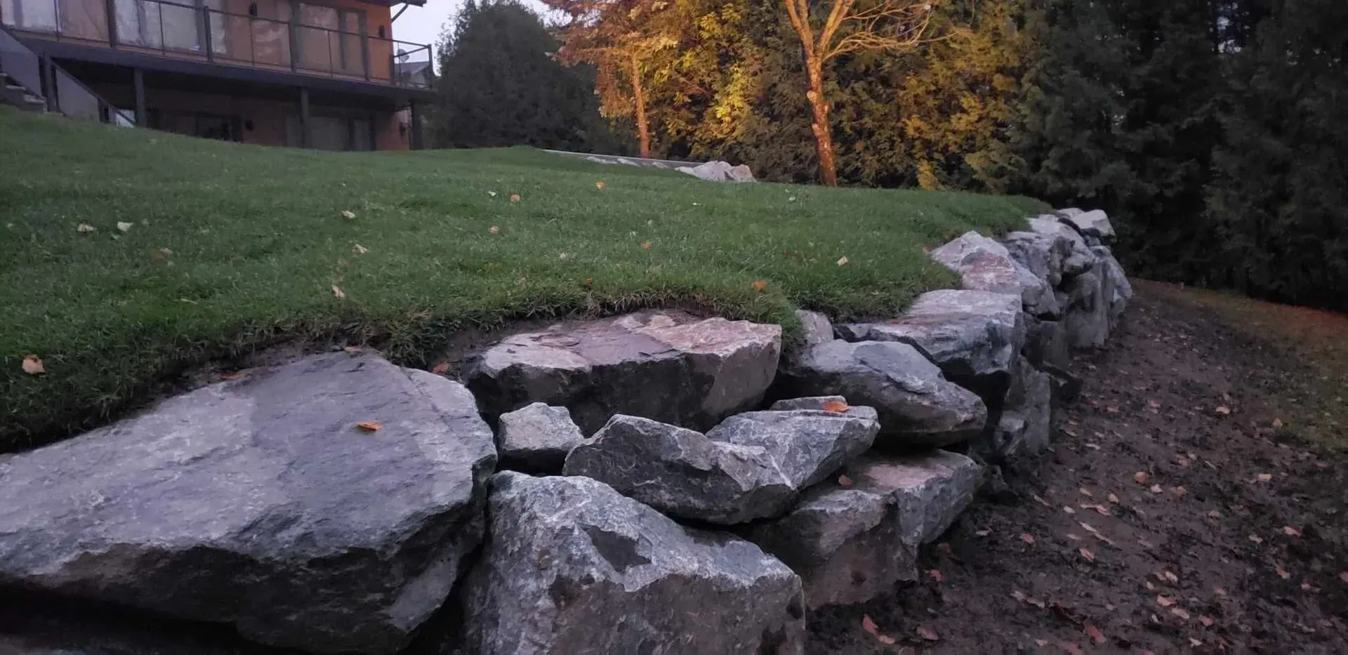 Mur de soutènement en pierre bordant une cour herbeuse au crépuscule, avec une maison et des arbres d'automne en arrière-plan.