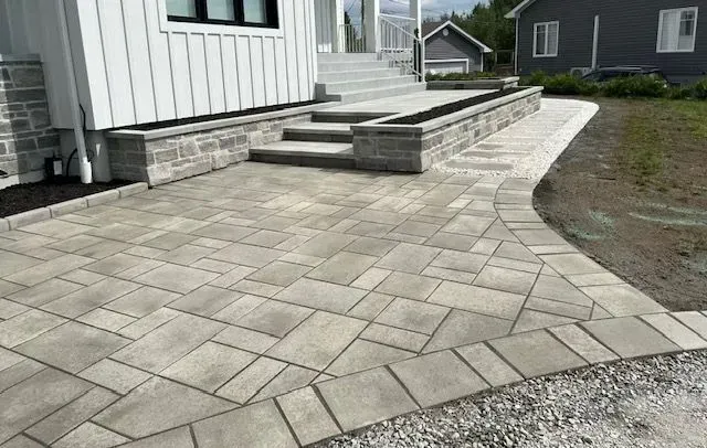 Terrasse en pavés de pierre jouxtant une maison, avec des marches et un bord arrondi.