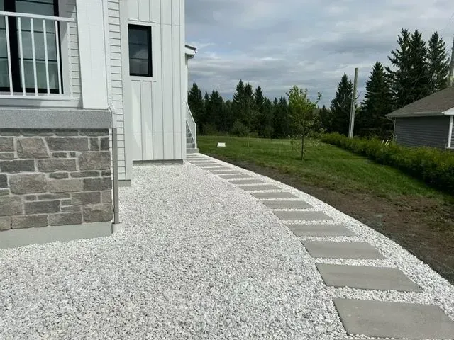 Allée de gravier longeant une maison blanche aux fondations en pierre et bordée de dalles de béton.