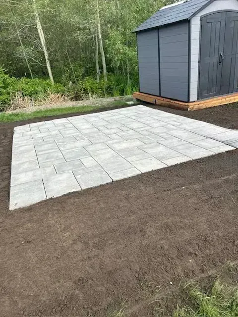 Terrasse en pierre grise jouxtant un hangar sombre sur un terrain en terre battue, avec des arbres en arrière-plan.