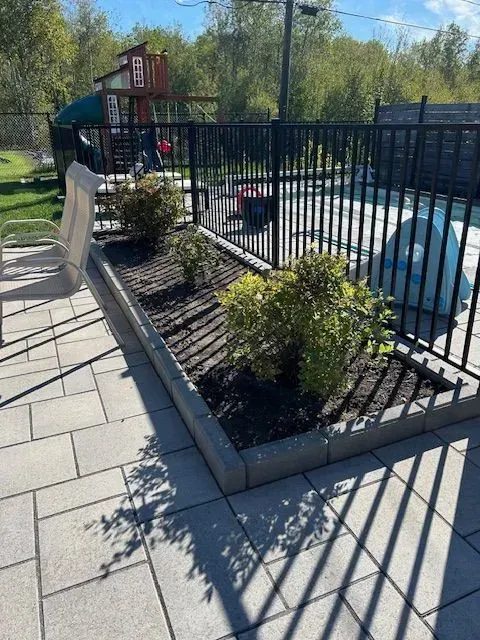 Jardinière surélevée le long d'une clôture en métal noir, avec des arbustes, une allée pavée et un objet recouvert d'une bâche bleue à proximité.