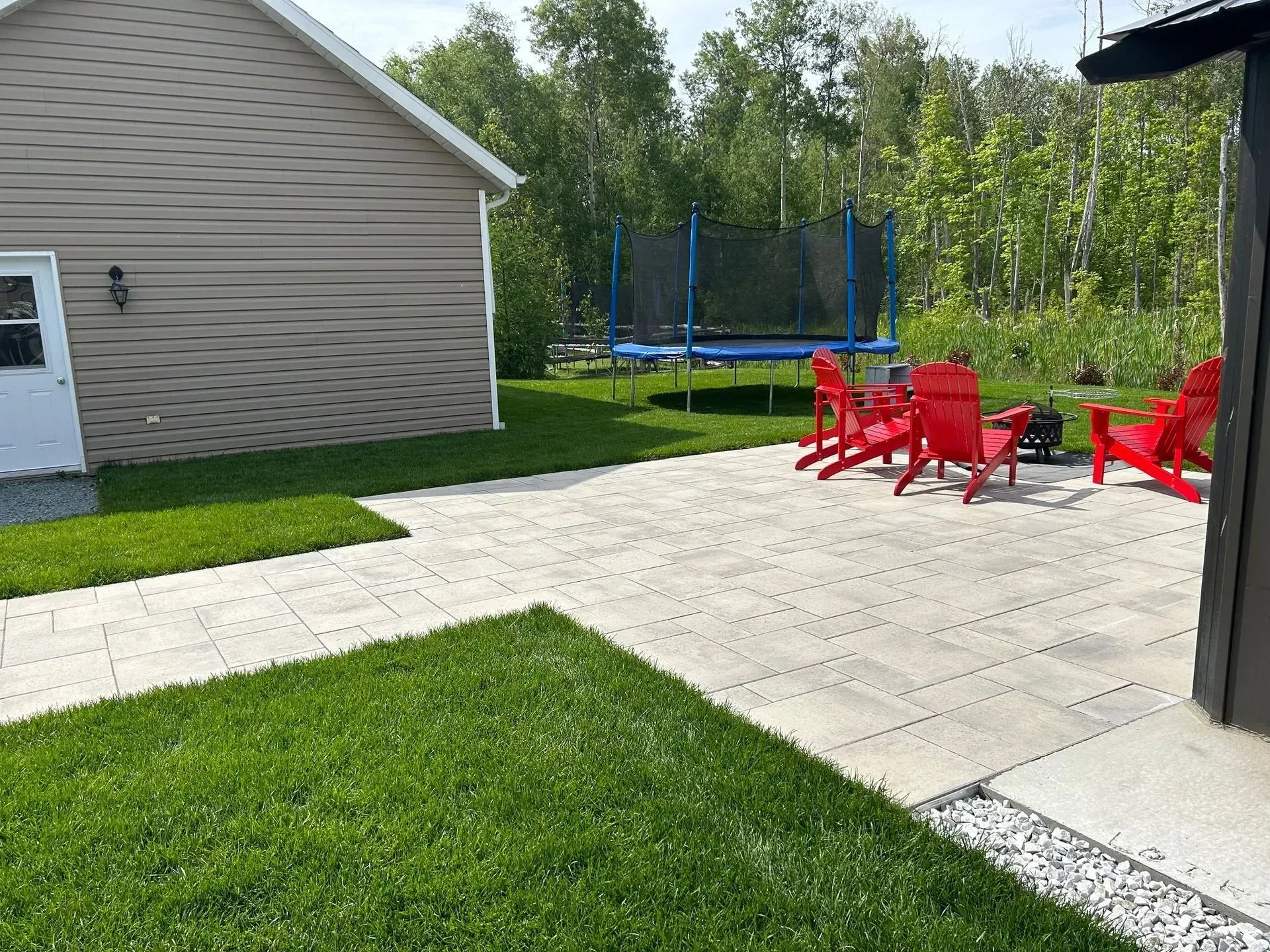 Terrasse arrière avec chaises rouges, dalles de béton, pelouse verte et trampoline près d'une maison