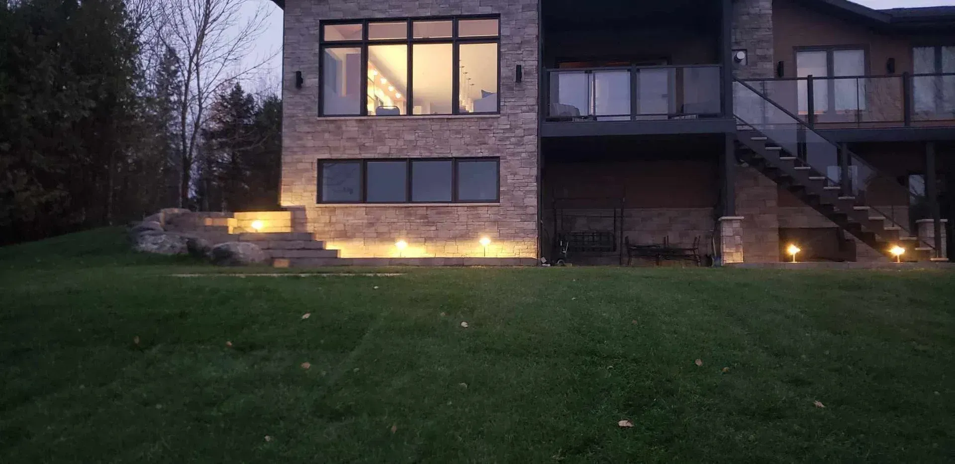 Maison moderne au crépuscule, fenêtres et balcon éclairés, donnant sur une pelouse vert foncé.