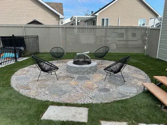 Terrasse arrière avec foyer circulaire en pierre et quatre chaises noires sur la pelouse.