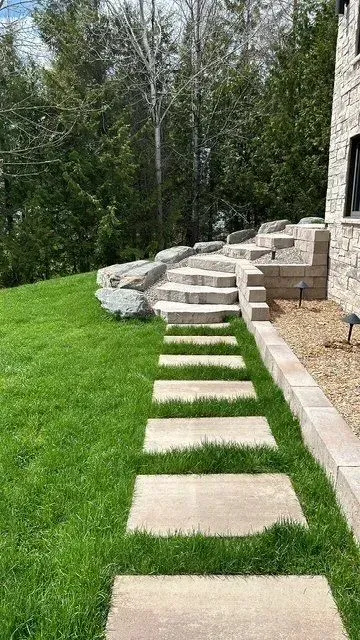 Un chemin de dalles traverse une pelouse verdoyante et mène à une terrasse en pierre jouxtant une maison et des arbres.
