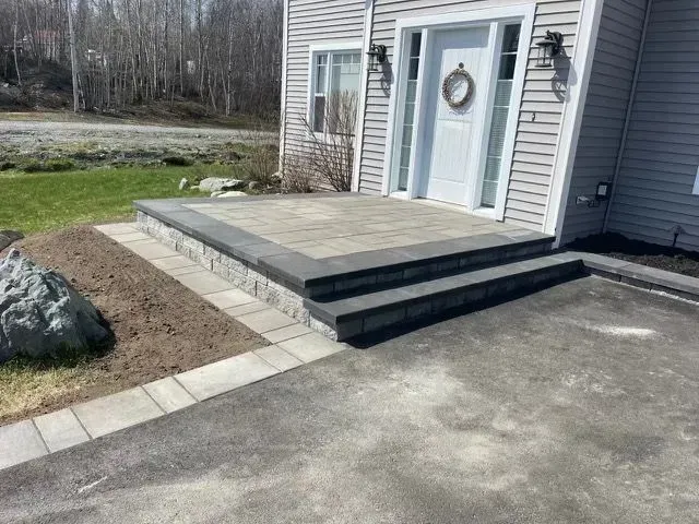 Entrée de la maison avec terrasse en béton à deux marches et allée longeant une pelouse et un gros rocher