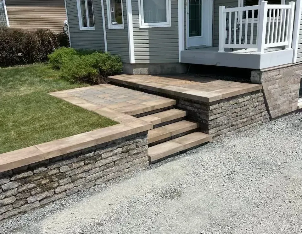 Terrasse en pierre avec marches et mur de soutènement menant au porche d'une maison, à côté d'une allée de gravier.